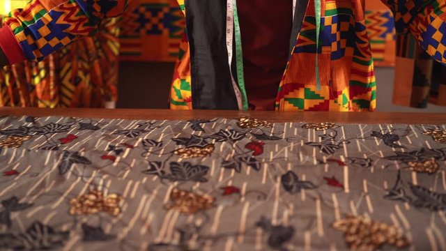 African American tailor in orange jacket cutting blue textile with scissors while working in sewing workshop on daytime