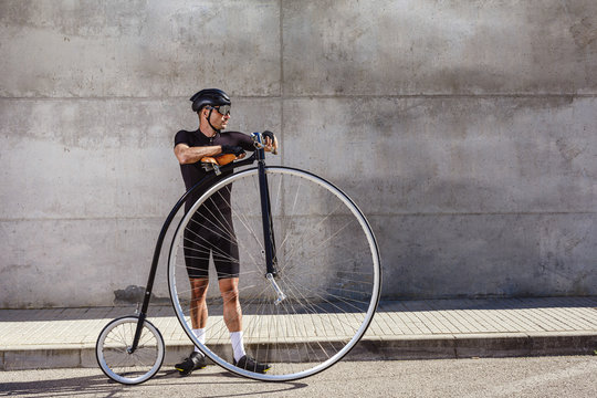 Calm determined man in black activewear and helmet looking away while standing on asphalt road and leaning on high wheel bicycle against concrete wall in sunny day