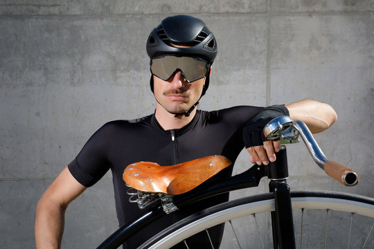 Calm determined man in black activewear and helmet looking at camera while standing on asphalt road and leaning on high wheel bicycle against concrete wall in sunny day