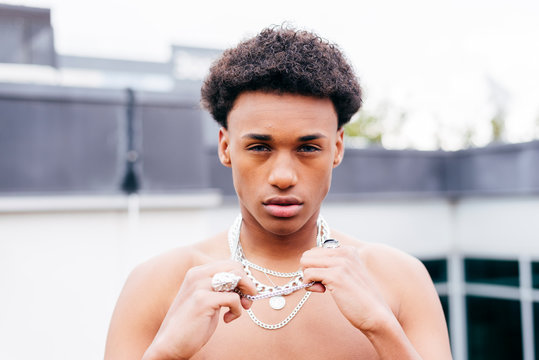 From above serious young African American male with naked torso wearing various trendy steel neck chains and pendant looking at camera while standing against modern building