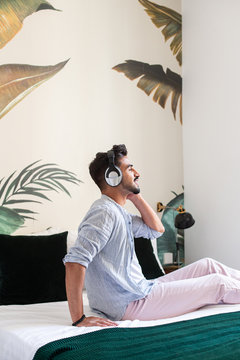 Side view of thoughtful bearded Hispanic male in headphones sitting on bed and enjoying good music at home
