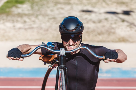 Portrait of happy man in black activewear and helmet looking at camera while standing with retro penny farthing bicycle on racetrack at sports ground
