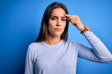 Young beautiful brunette woman wearing casual sweater standing over blue background pointing unhappy to pimple on forehead, ugly infection of blackhead. Acne and skin problem
