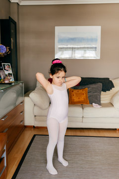 Cute little girl in light pink leotard and tights getting ready while standing near dance shoes in cozy living room at home