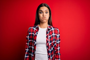 Young beautiful brunette woman wearing casual shirt standing over isolated red background puffing cheeks with funny face. Mouth inflated with air, crazy expression.