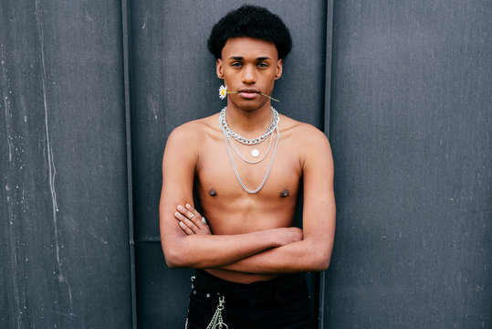 Alluring young black teenage man with naked torso and neck chains holding flower in mouth and looking at camera while standing against gray wall