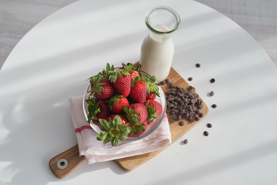 From above bowl of ripe strawberries and heap of chocolate pellets placed on napkin and cutting board near bottle of milk on kitchen table in morning