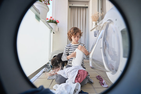 Playful boy in casual clothes having fun with clothes while sitting with dog in front of washing machine in laundry room