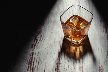 From above transparent square shaped glass of whiskey drink with ice cubes on shabby wooden table in rays in dark room