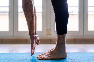 Low angle side view of unrecognizable flexible barefoot man in sportswear doing standing forward bend exercise while training alone on sports mat against window in light spacious room at home