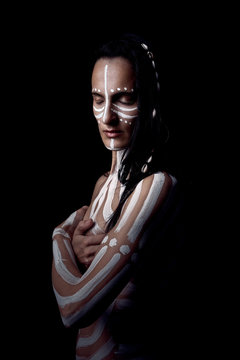 Side view portrait of naked tender beautiful brunette Native American woman with white striped painted on body covering breast standing in dark on black background with closed eyes