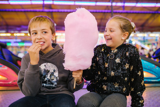 Delighted girl and boy siblings smiling and eating sweet candyfloss while sitting at funfair