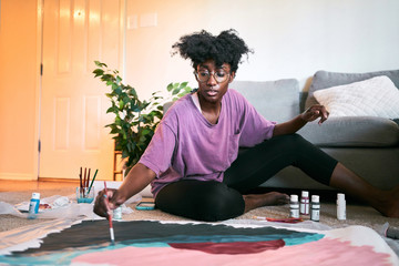 Young happy beautiful African American woman, amateur artist in casual outfit drawing colorful picture with paintbrush on paper while sitting on floor in cozy apartment