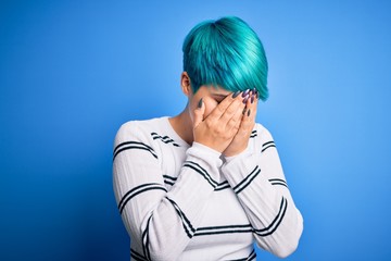 Young beautiful woman with blue fashion hair wearing casual sweater standing at studio with sad expression covering face with hands while crying. Depression concept.