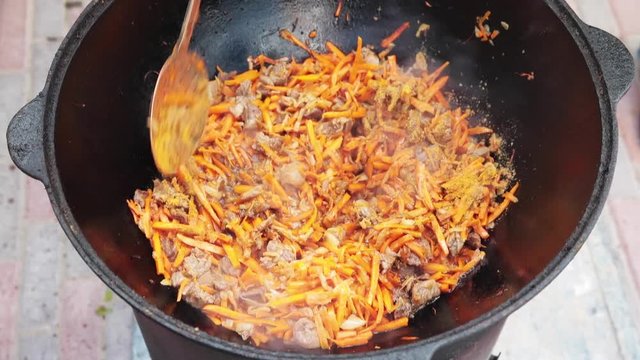 Preparation of national uzbek dish pilaf, pilaw, plov, carrot with meat in big pan. Cooking process, open fire. Cooking in a cauldron on fire. Stir slowly with a skimmer. Add spices