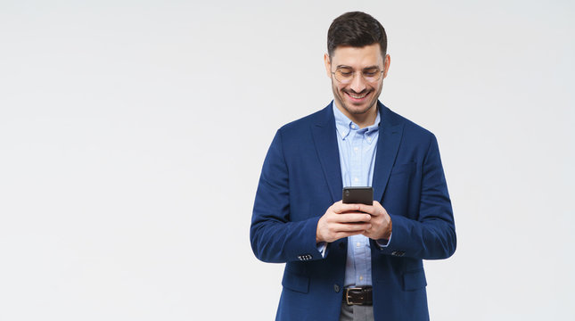 Horizontal banner of young man in glasses, blue blazer and shirt, smiling at content on his smart phone screen, copy space on left, isolated on gray background
