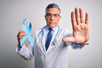 Middle age handsome grey-haired doctor man wearing coat holding blue cancer ribbon with open hand doing stop sign with serious and confident expression, defense gesture