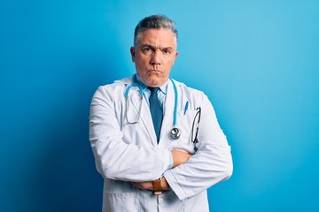 Middle age handsome grey-haired doctor man wearing coat and blue stethoscope skeptic and nervous, disapproving expression on face with crossed arms. Negative person.