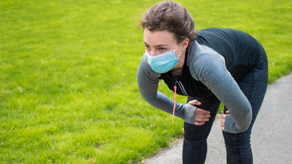 Sport during quarantine, coronavirus, covid-19 concept. Tired young athletic woman runner wearing medical protective mask taking a rest after running hard outdoors. Sterilizing face mask protection