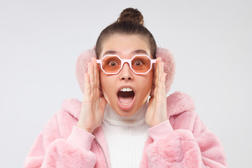 Wow! Winter photo of young woman wearing pink fluffy coat, ear warmers and eyeglasses, feeling...