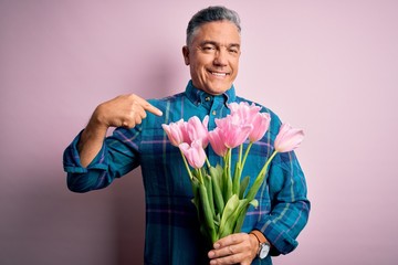 Middle age grey-haired man holding natural bouquet of pink tulips over isolated background with surprise face pointing finger to himself