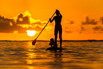 Young mother with a small child ride a S.U.P. (paddle) board in the Indian Ocean on the background of an incredible sunset