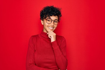 Fototapeta na wymiar Young beautiful african american afro woman wearing turtleneck sweater and glasses looking confident at the camera smiling with crossed arms and hand raised on chin. Thinking positive.