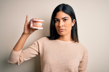 Young beautiful hispanic dentist woman holding prosthesis dental mouth with a confident expression on smart face thinking serious