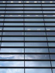 a vertical perspective view of the facade of a modern glass commercial building with steel frames with clouds and sky reflected in the windows