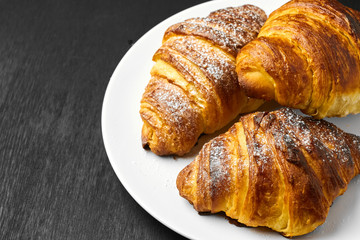 Top view on three fresh french buttery croissant on a white plate on a black background with copy space