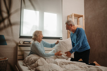 Obraz premium Happy elderly couple having a pillow fight on bed in their bedroom.