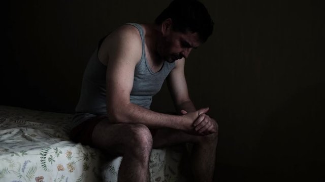 Tired pensive depressed man sitting on a bed in a dark room	

