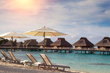 chairs on the sandy beach and houses over water of the blue sea. Polynesia.