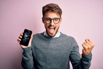 Young man with beard wearing glasses holding broken and craked smartphone screaming proud and...
