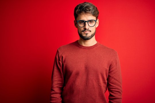 Young handsome man with beard wearing glasses and sweater standing over red background Relaxed with serious expression on face. Simple and natural looking at the camera.