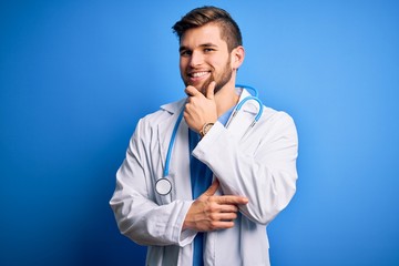 Young blond doctor man with beard and blue eyes wearing white coat and stethoscope looking confident at the camera with smile with crossed arms and hand raised on chin. Thinking positive.