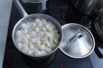 Ravioli in pan, boiling water, silver metal background. Cooking Pelmeni, russian dumplings stuffed with minced meat, in steel stewpot on ceramic stove in home kitchen. Varenyky, vareniki.