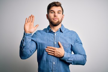 Young handsome blond man with beard and blue eyes wearing casual denim shirt Swearing with hand on...