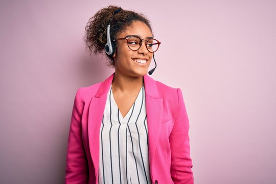 Young african american call center agent girl wearing glasses working using headset looking away to side with smile on face, natural expression. Laughing confident.