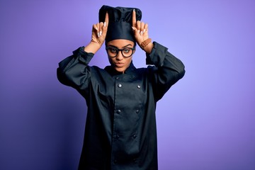 Young african american chef girl wearing cooker uniform and hat over purple background doing funny...