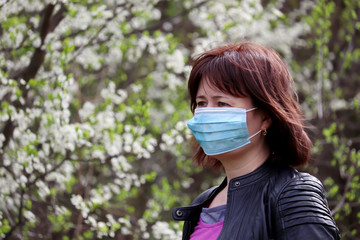 Woman in medical protective mask in a spring garden on cherry blossom background. Concept of quarantine during covid-19 coronavirus pandemic