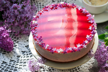 A berry mousse cake with chocolate base and juice jelly, decorated with lilac flowers, served with coffee.