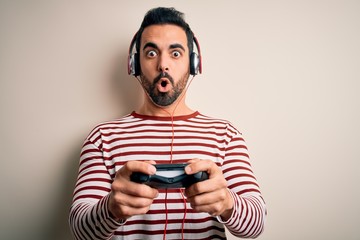 Young handsome gamer man with beard playing video game using joystick and headphones scared in...