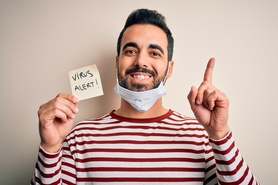 Young handsome man with beard wearing medical mask holding virus alert reminder surprised with an idea or question pointing finger with happy face, number one