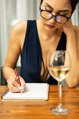 A woman wearing glasses sits at a table drinking white wine and writing in a journal