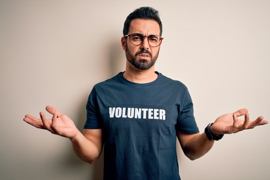 Handsome man with beard wearing t-shirt with volunteer message over white background clueless and confused with open arms, no idea concept.