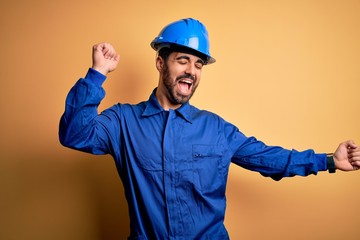 Mechanic man with beard wearing blue uniform and safety helmet over yellow background Dancing happy and cheerful, smiling moving casual and confident listening to music