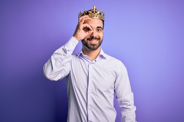 Young handsome man with beard wearing golden crown of king over purple background doing ok gesture with hand smiling, eye looking through fingers with happy face.