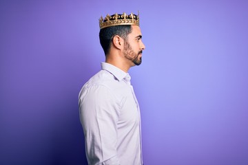 Young handsome man with beard wearing golden crown of king over purple background looking to side,...