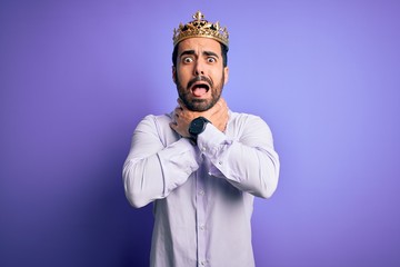 Young handsome man with beard wearing golden crown of king over purple background shouting and suffocate because painful strangle. Health problem. Asphyxiate and suicide concept.